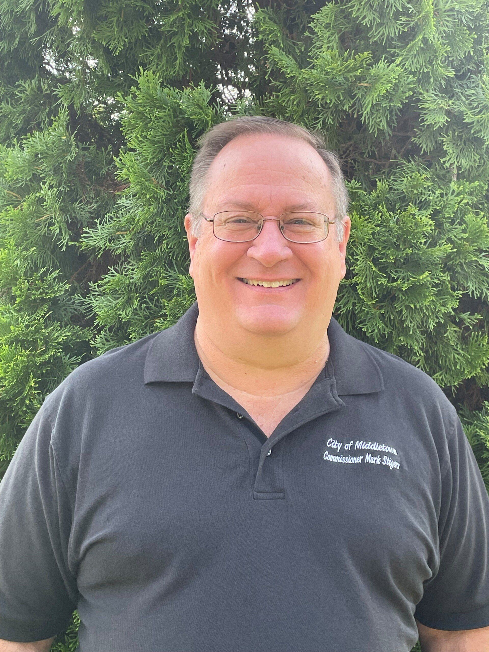 A person in a dark polo shirt stands in front of a green bush, smiling at the camera.