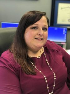 A person sits at a desk wearing a purple top and necklace, with computer monitors and a framed certificate in the background.