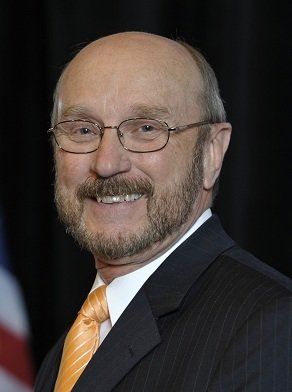 A man in a suit and orange tie smiles in front of a dark background with a partial view of a flag.