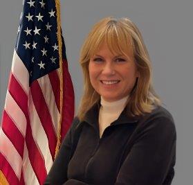 'Portrait of a woman with shoulder-length blonde hair, smiling and standing in front of the American flag. She is wearing a dark jacket over a white turtleneck. Neutral gray background.'