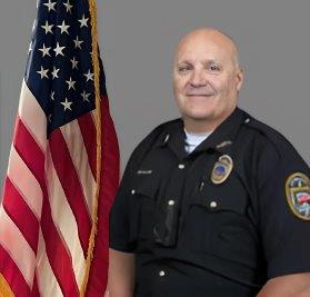 Portrait of a man in a black police uniform with badge, shoulder patch, and a radio. He stands in front of the American flag and is smiling slightly. Neutral gray background.