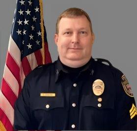 Portrait of a man with short hair in a dark police uniform with a badge, name tag, and sergeant stripes on his sleeve. He is standing in front of the American flag and has a neutral expression. Gray background.
