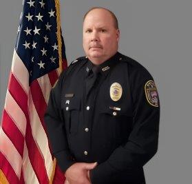 Portrait of a man with light brown hair in a black police uniform with badge and shoulder patch, standing with hands folded in front of the American flag. He has a serious expression. Gray background.